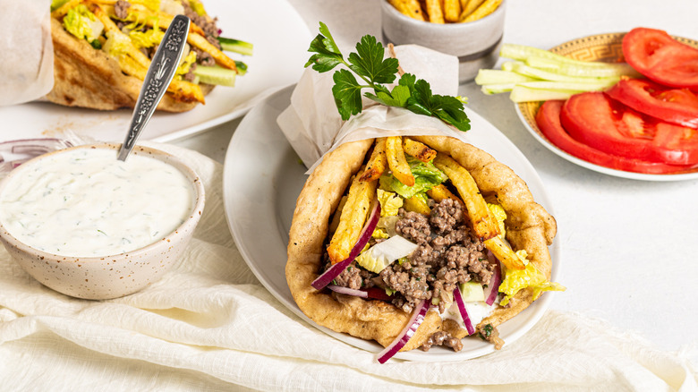 Ground beef gyros on a plate with a side of tzatziki and a side of sliced tomatoes and cucumbers