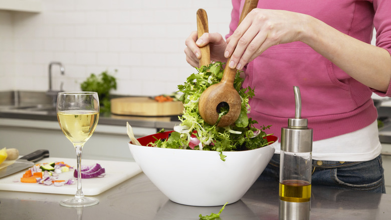 A glass of wine next to a bowl of salad being tossed