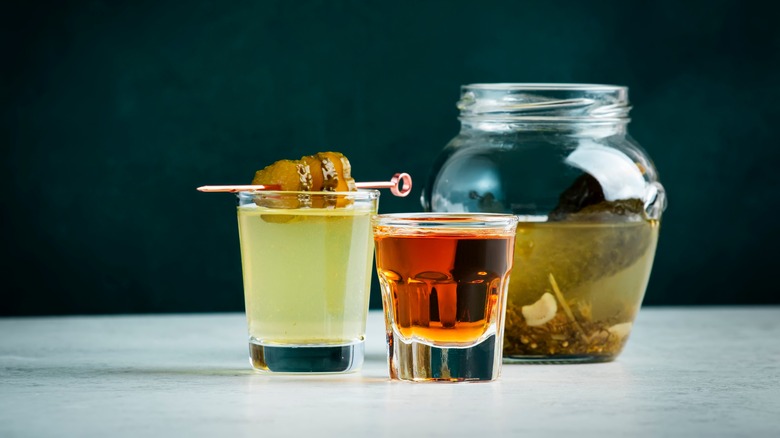 A shot of whiskey served with a shot of pickle juice, with a jar of cucumber pickles in the background