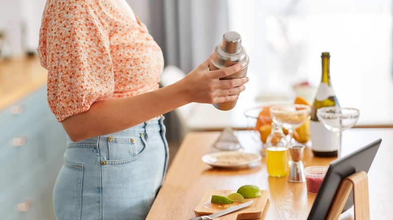 Person holding cocktail shaker in home kitchen with cocktail ingredients