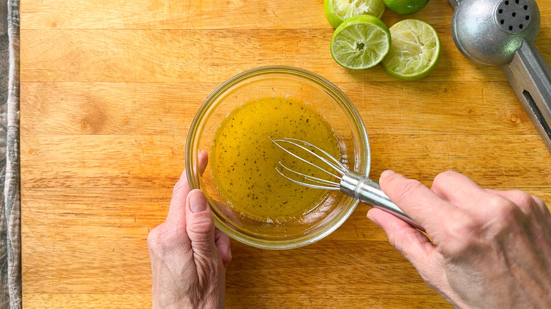 Whisking dressing for deconstructed watermelon gazpacho salad in glass bowl on cutting board with lime halves and juicer