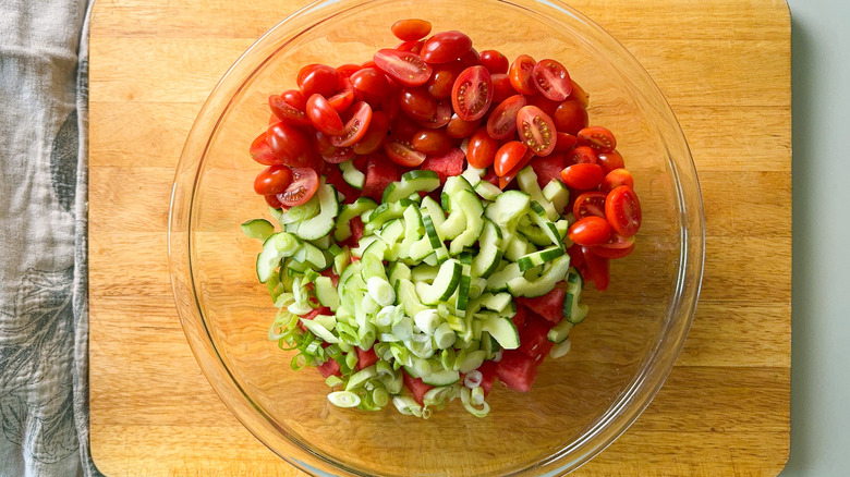 Cut watermelon, cucumber, grape tomatoes, and scallions in glass bowl on cutting board